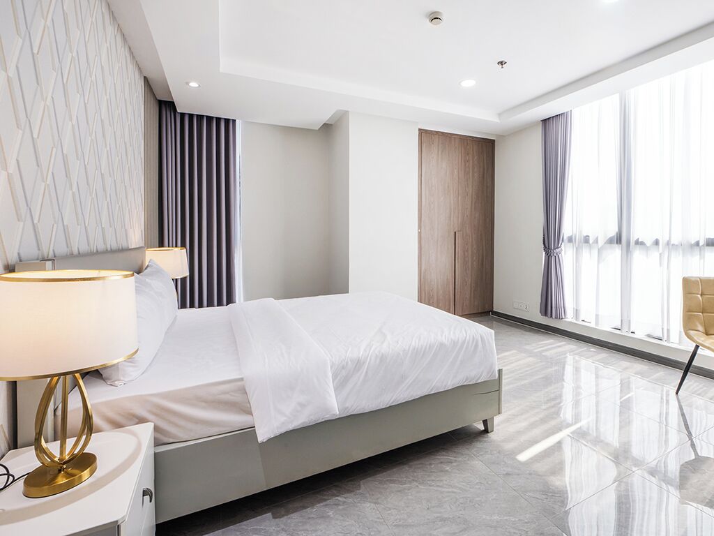 A modern hotel room with a bed, a nightstand, a lamp, a chair, and large windows.