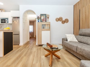 Ein modernes Wohnzimmer mit einem Glastisch, einem grauen Sofa und einer Küchenzeile inklusive Mikrowelle.