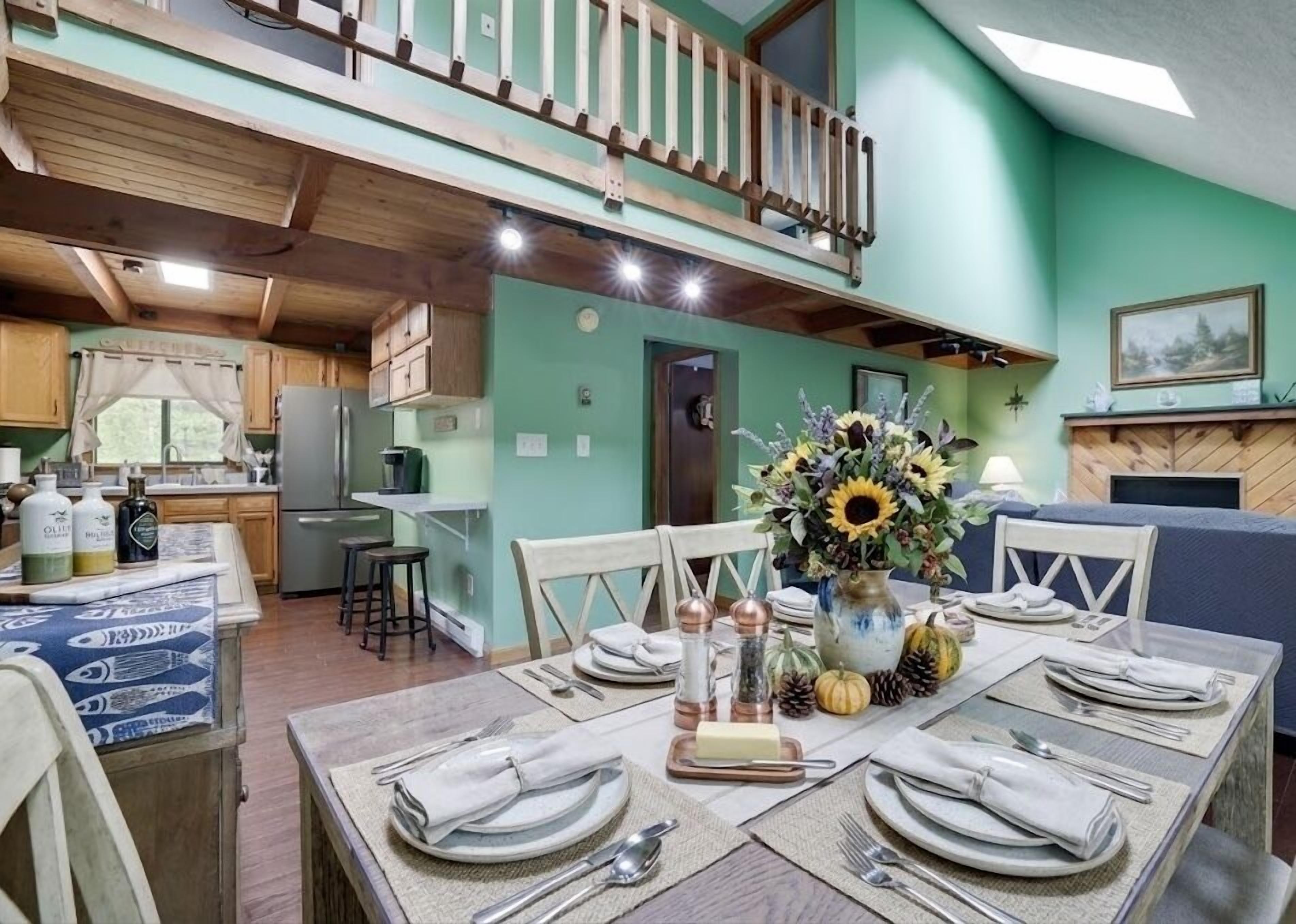 A dining area with a table set for four, a kitchen in the background, and a balcony with a wooden railing.