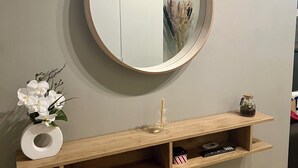 Ein an der Wand befestigter Spiegel mit Holzrahmen, darunter ein Holzregal und eine Vase mit weißen Blumen.