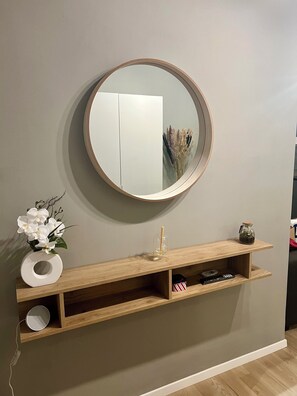 Ein an der Wand befestigter Spiegel mit Holzrahmen, darunter ein Holzregal und eine Vase mit weißen Blumen.