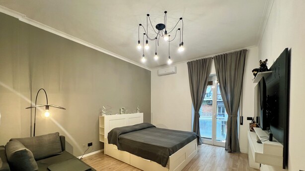 Ein modernes Schlafzimmer mit Bett, Schreibtisch, Stuhl, Fernseher und einem Balkon mit Vorhängen.