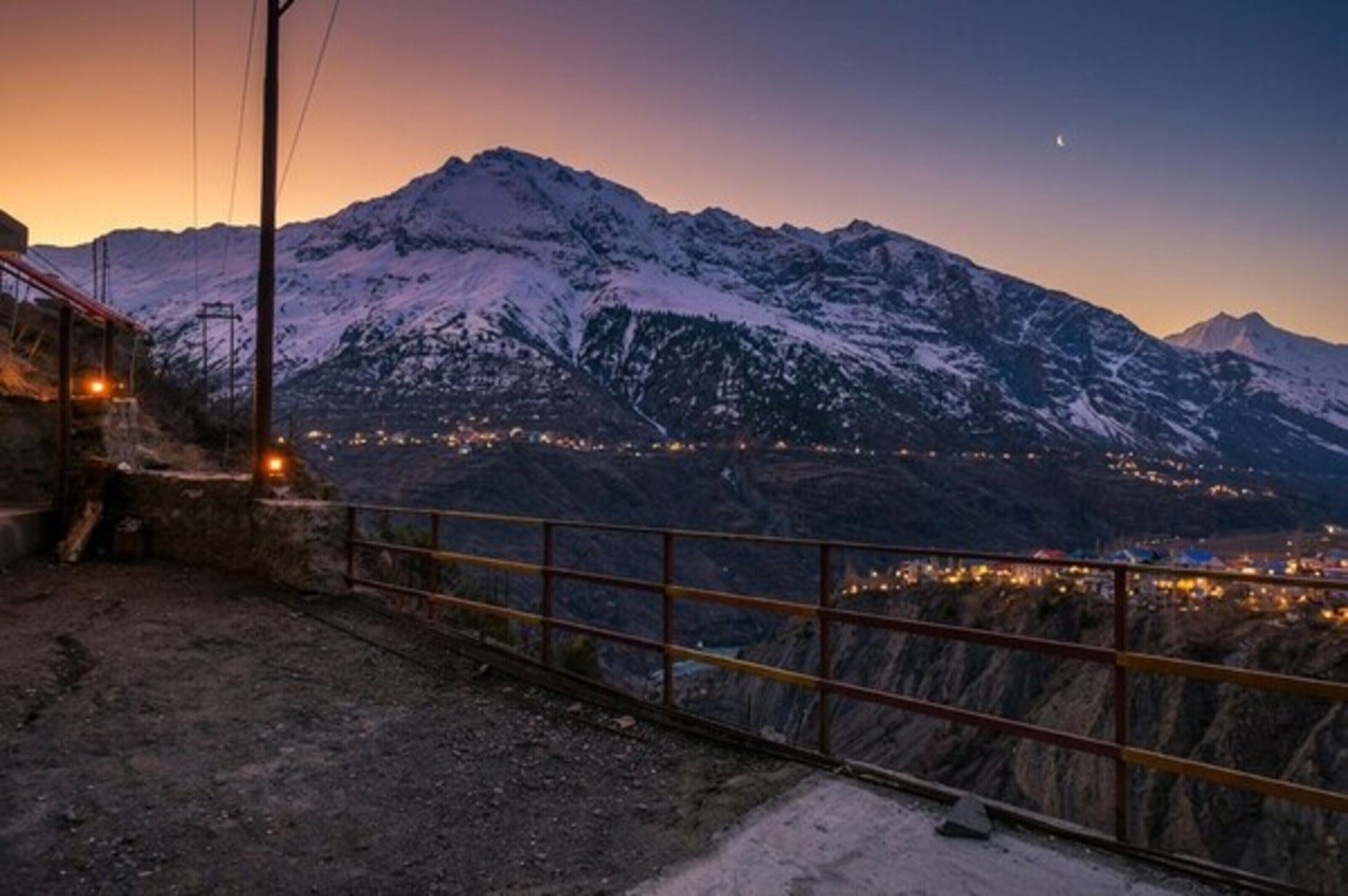 A mountainous landscape with a snow-covered peak, a distant town, and a metal railing.