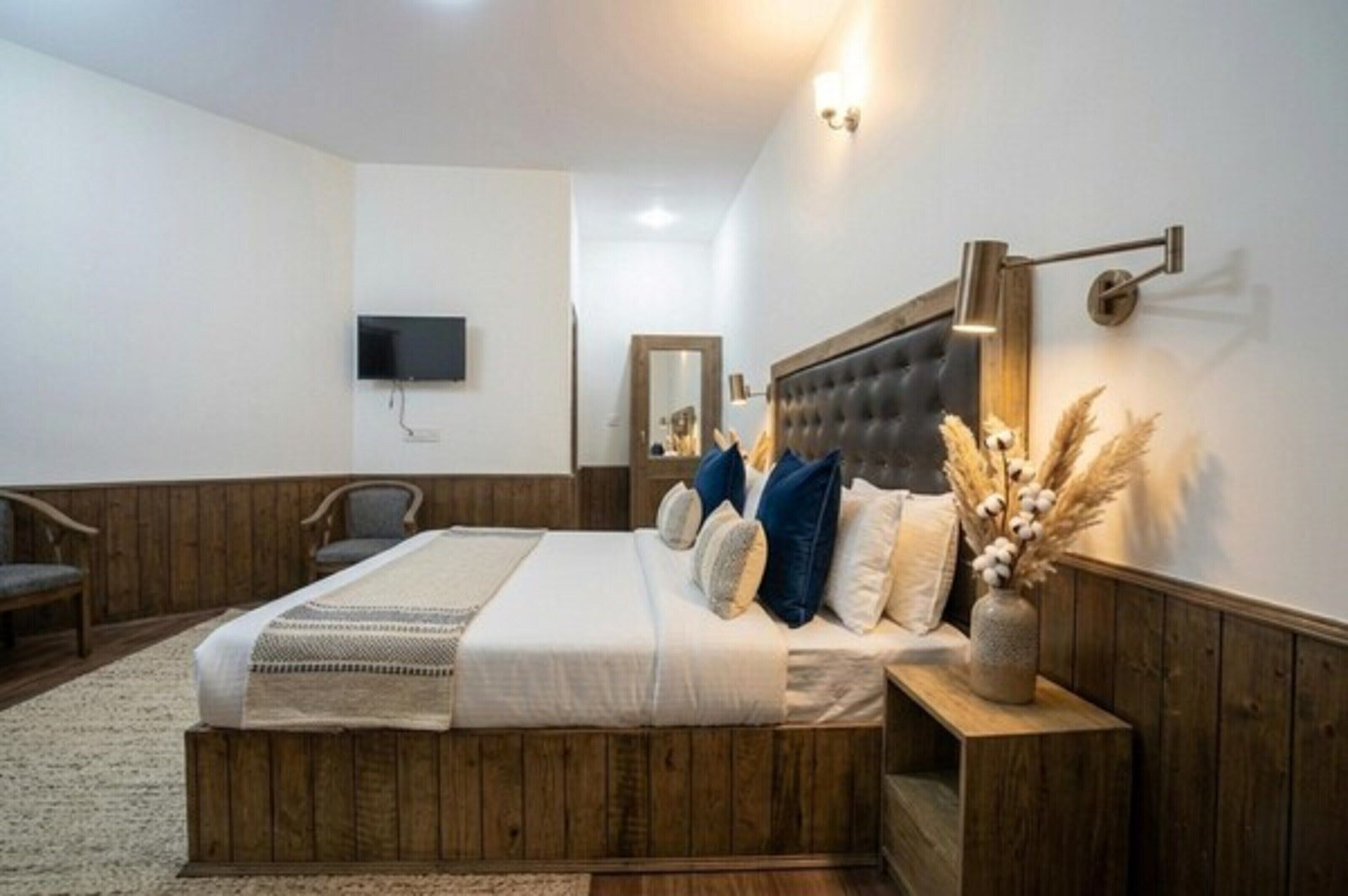 A bedroom with a wooden bed frame, a flat-screen TV mounted on the wall, and a bedside table with a vase of dried flowers.
