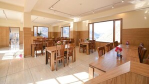 A spacious dining area with multiple wooden tables and chairs, large windows with mountain views, and a modern interior design. - Biker Gang Hotel & Cafe By OTABoostup (Keylong)