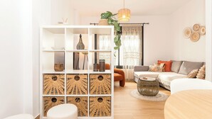 Ein modernes Wohnzimmer mit einem weißen Sofa, einem runden Couchtisch, einem weißen Hocker, einem Bücherregal mit gewebten Körben und einer hängenden Pflanze.
