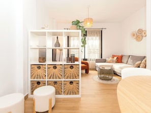 Ein modernes Wohnzimmer mit einem weißen Sofa, einem runden Couchtisch, einem weißen Hocker, einem Bücherregal mit gewebten Körben und einer hängenden Pflanze.
