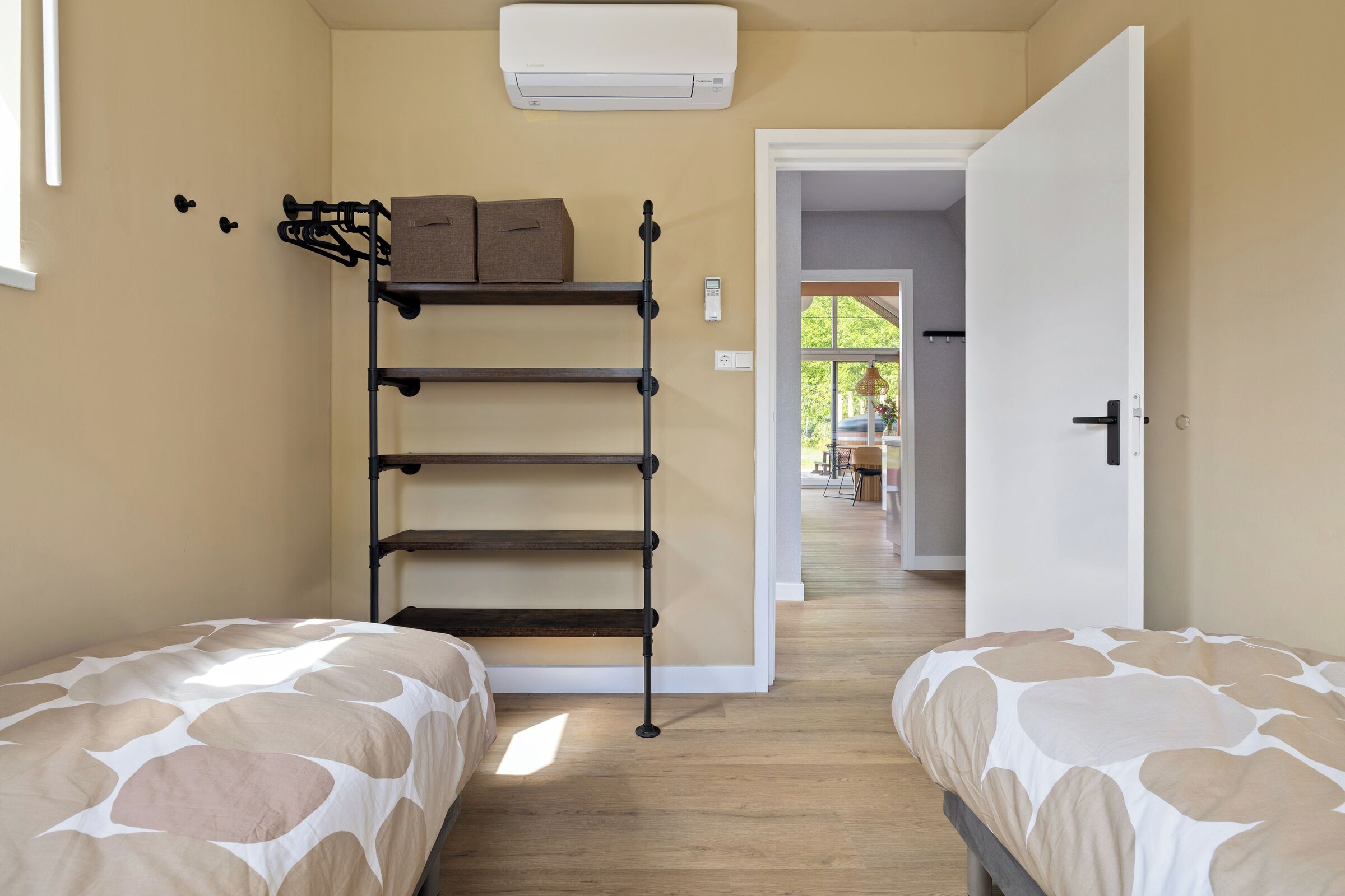 Een slaapkamer met twee bedden, een inbouwplank met dozen, een airconditioning en een deur die naar een andere kamer leidt.
