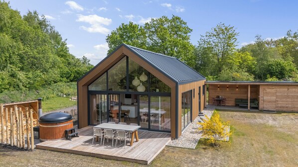 Una cabaña moderna de madera con frente de vidrio, una terraza amoblada y un jacuzzi.