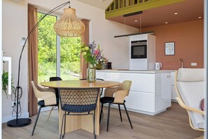 Una cocina moderna con armarios blancos, un comedor con una mesa redonda y sillas, y un ventanal con vistas a zonas verdes.