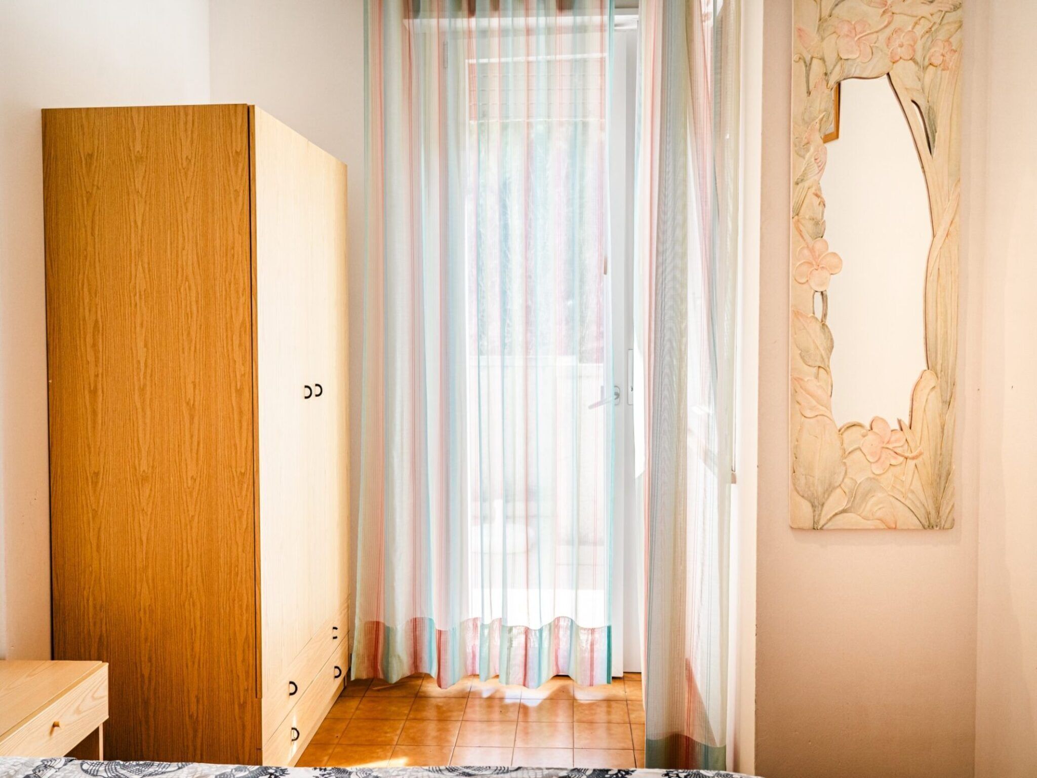 A room with a wooden wardrobe, a bed with a patterned bedspread, a window with sheer curtains, and a wall with a floral design.