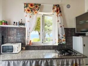 Eine Küche mit Mikrowelle, Gaskochfeld und Meerblick durch ein Fenster mit floralen Vorhängen.