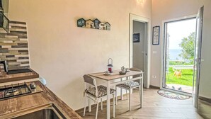 Cozinha com fogão a gás, mesa de jantar com cadeiras, um refrigerador pequeno e vista para o exterior por uma porta de vidro.