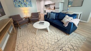 A living room with a blue sofa, a coffee table, and a patterned rug. There is a painting on the wall and a kitchen area in the background. - Twin Oaks Extended Stay One Bedroom - 2817 Apt 13 (Syracuse)