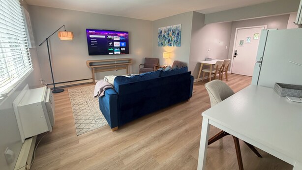 A modern living room with a blue sofa, a flat-screen TV, a dining table, and a kitchen area. - Twin Oaks Extended Stay One Bedroom - 2817 Apt 13 (Syracuse)
