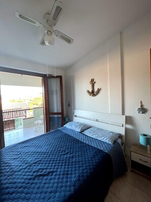 A bedroom with a bed, a ceiling fan, a balcony, and a wall-mounted anchor decoration. - Small villa in quiet setting a few minutes from the sea (Villasimius)