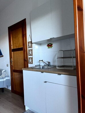 A compact kitchen with a wooden door, white cabinets, a stainless steel sink, and a small fruit decoration. - Small villa in quiet setting a few minutes from the sea (Villasimius)