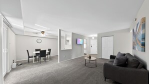 Uma sala moderna com um sofá cinzento, uma área de refeições com mesa e cadeiras, uma TV fixada na parede e um ventilador de teto.