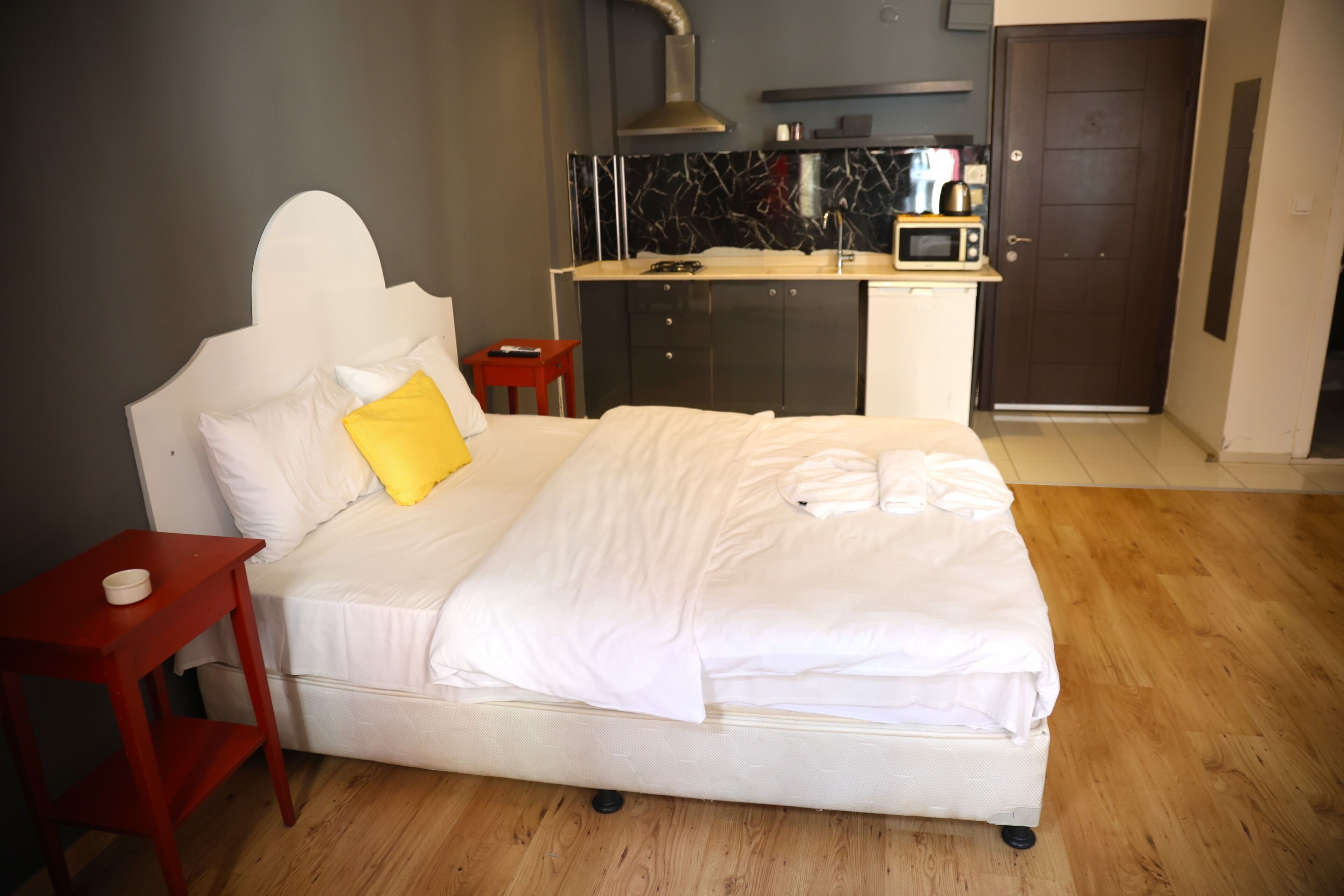 Ein Hotelzimmer mit einem Bett, einem kleinen Nachttisch in Rot, Holzboden, einer Küchenzeile mit einer Mikrowelle und einer Tür.