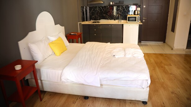 A hotel room with a bed, a small red nightstand, a wooden floor, a kitchenette with a microwave, and a door. - BRK DREAM SUITS (Istanbul)