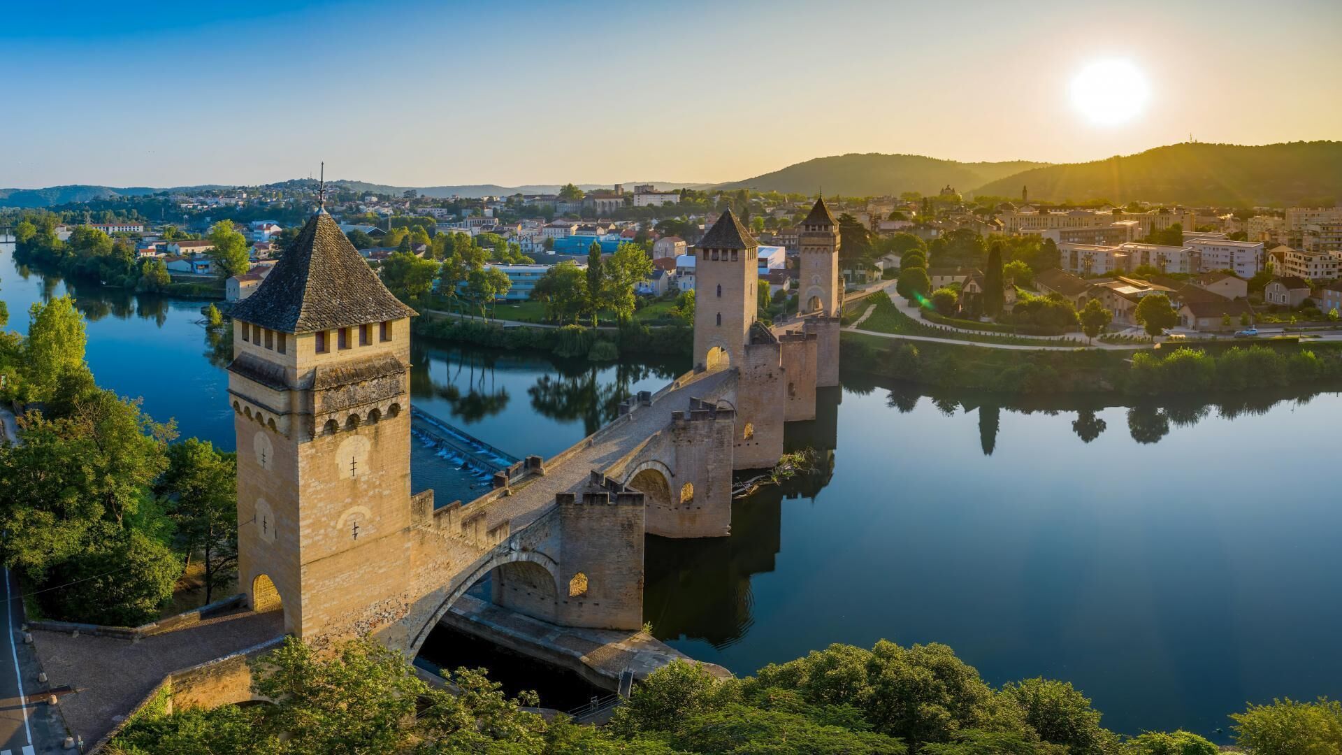 A historic stone bridge with towers over a calm river, surrounded by greenery and a town in the background.