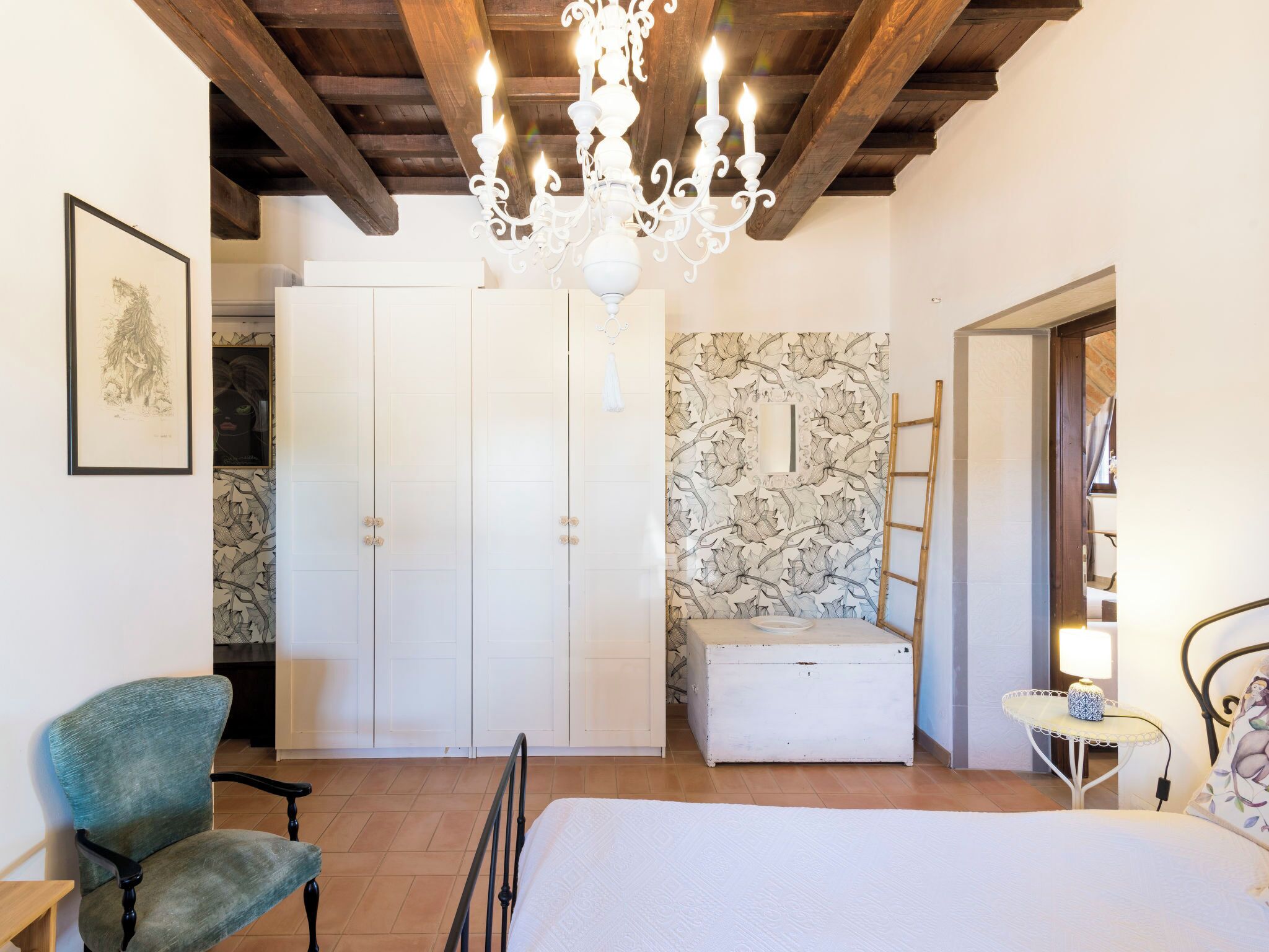 Une chambre à coucher comprenant un lit, une chaise, une commode, une armoire, un lustre et un mur orné d’un motif de feuilles.