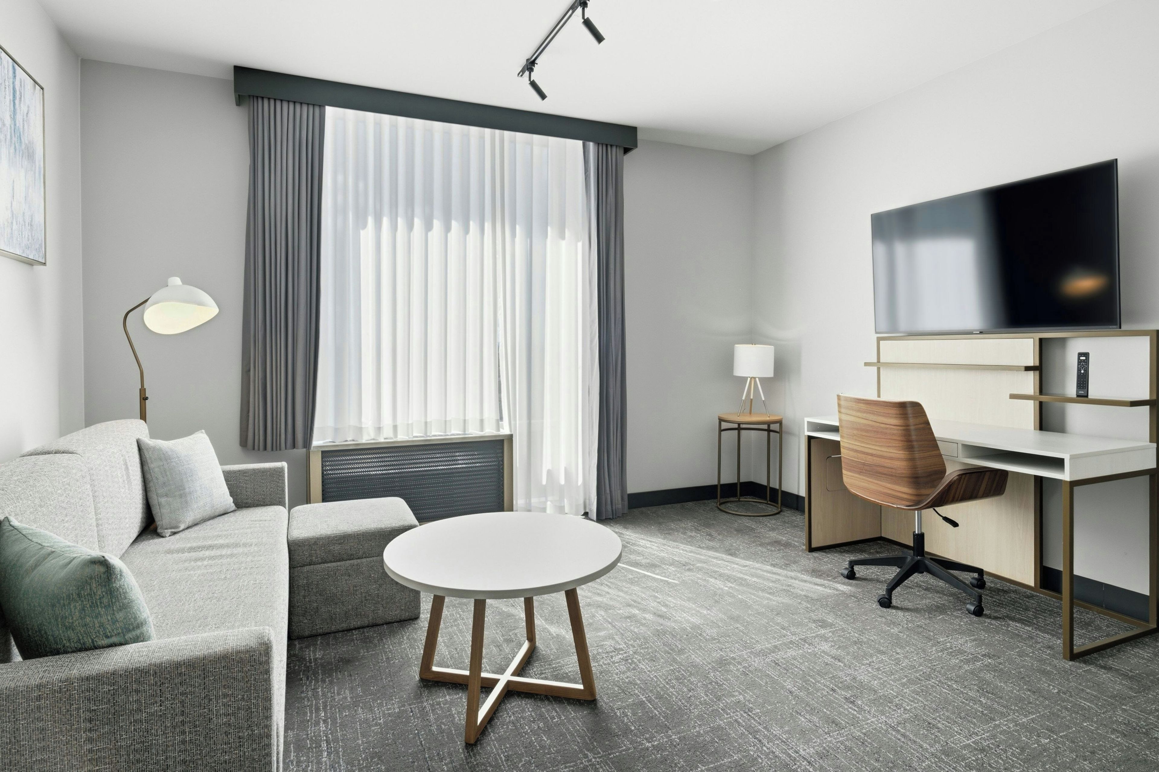 Een moderne hotelkamer met een grijze bank, een ronde tafel, een bureau met stoel, een televisie en een raam met gordijnen.