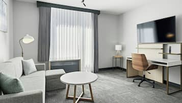 A modern hotel room with a grey sofa, a round table, a desk with a chair, a TV, and a window with curtains.
