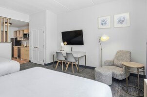 Una habitación de hotel con una cama, un escritorio con dos sillas, un televisor, una lámpara, una mesita pequeña y un sofá.