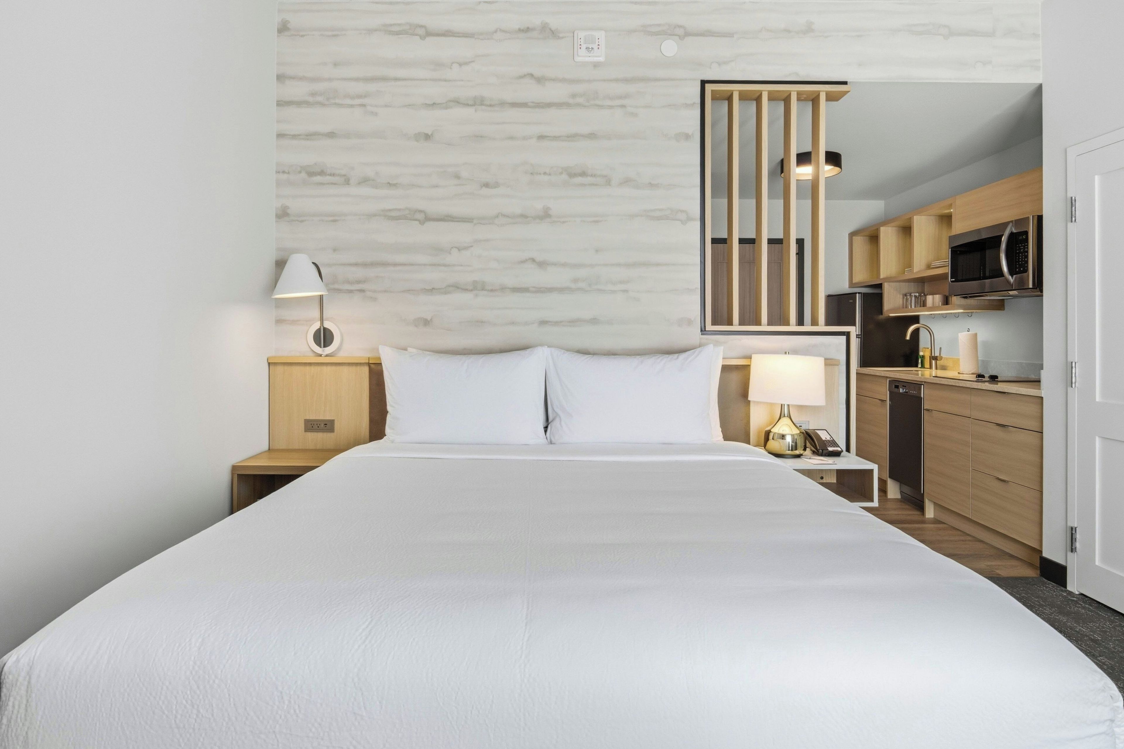 A hotel room with a large bed, a bedside table with a lamp, a small desk with a lamp, and a kitchenette with a microwave and refrigerator.