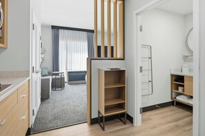 A modern hotel room with a kitchenette, a seating area with a sofa and a small table, a wooden shelf unit, and a bathroom with a towel rack. - TownePlace Suites by Marriott Vaughan (Vaughan)