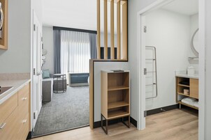 Una habitación de hotel moderna con kitchenette, una zona de estar con sofá y una mesa pequeña, un estante de madera y un baño con perchero.