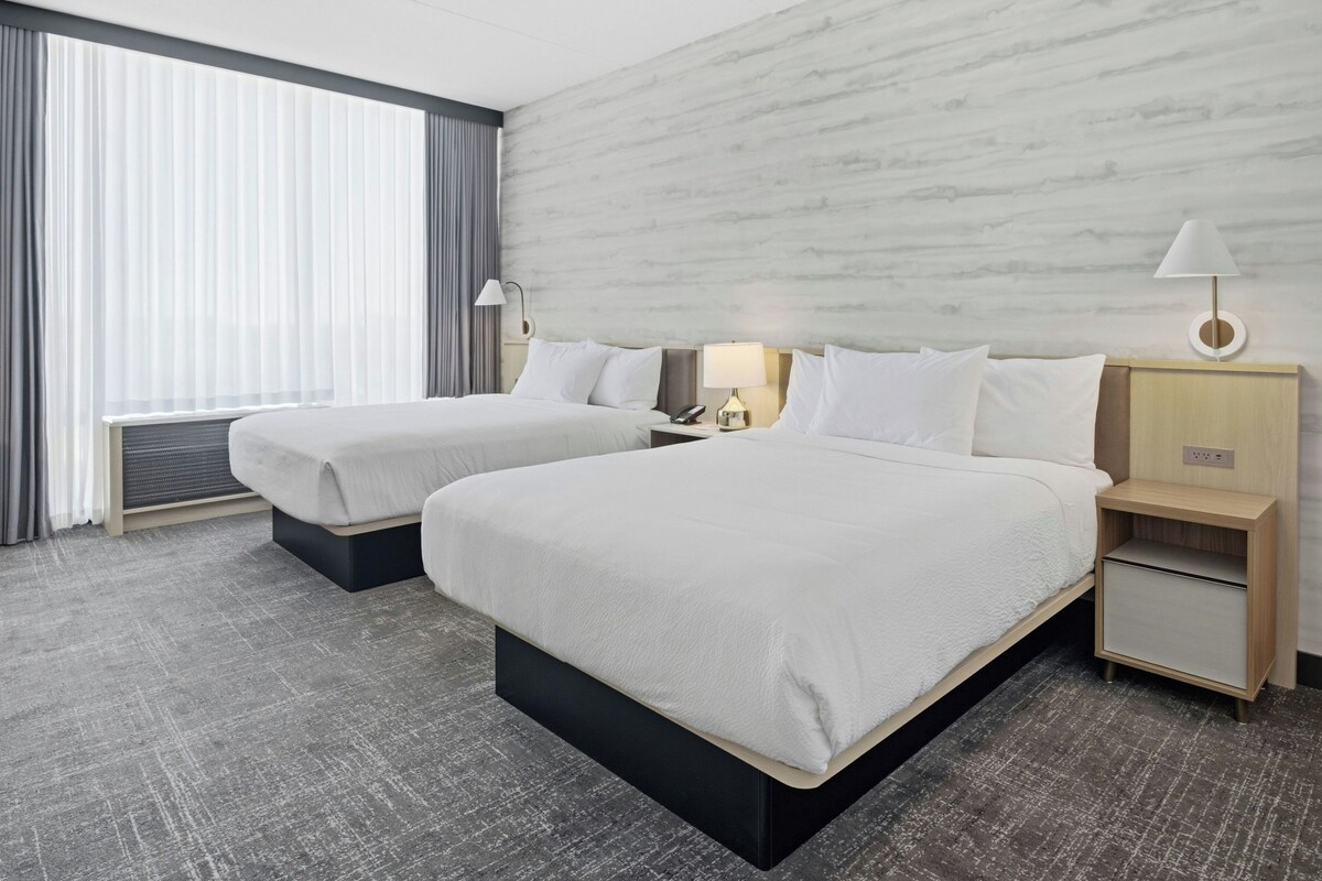 A hotel room with two beds, a nightstand, a lamp, and a view of the outdoors through the window.