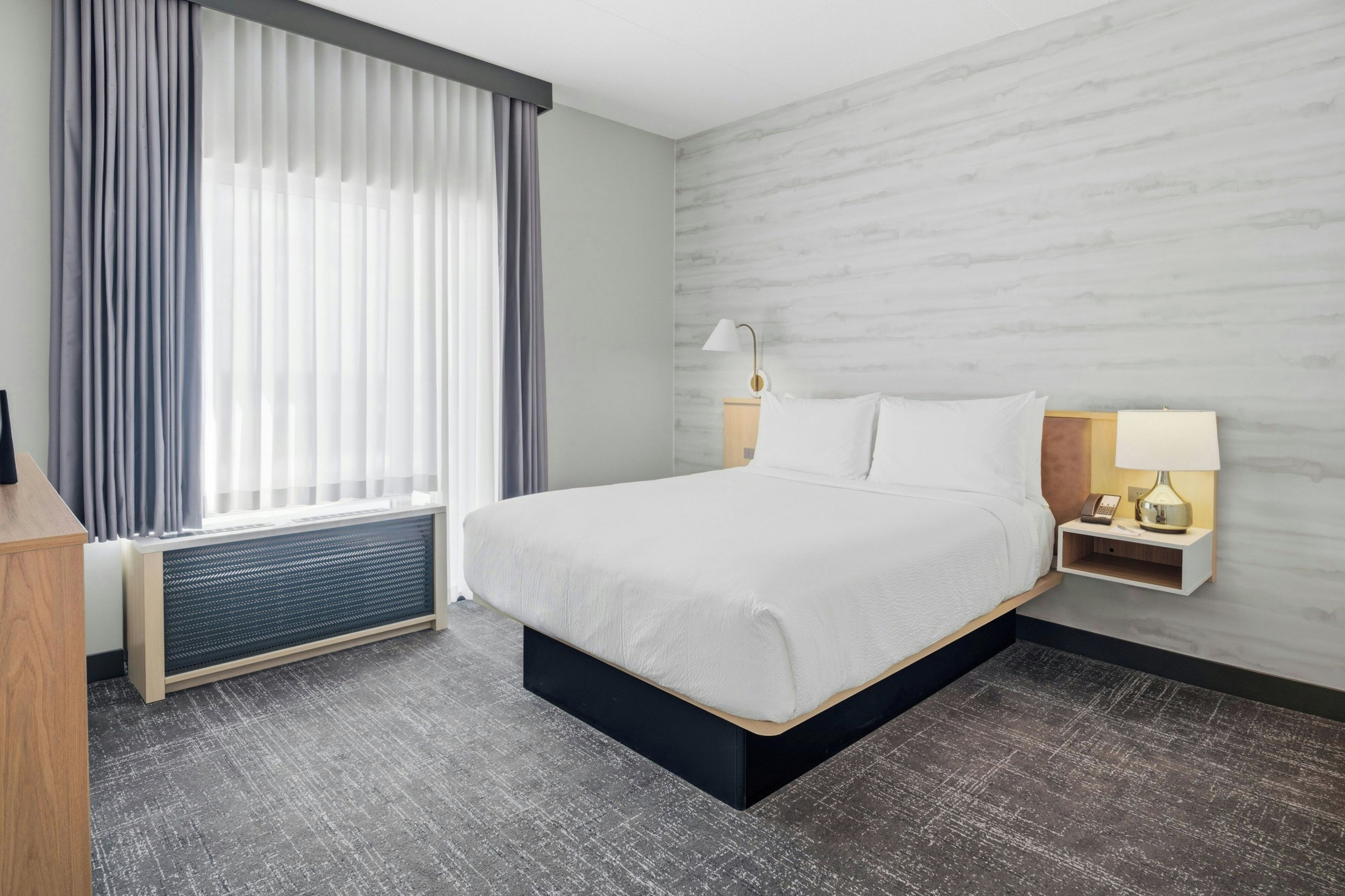 Ein Hotelzimmer mit einem Bett, einem Nachttisch mit Lampe, einem Fenster mit Vorhängen und einem Heizkörper.