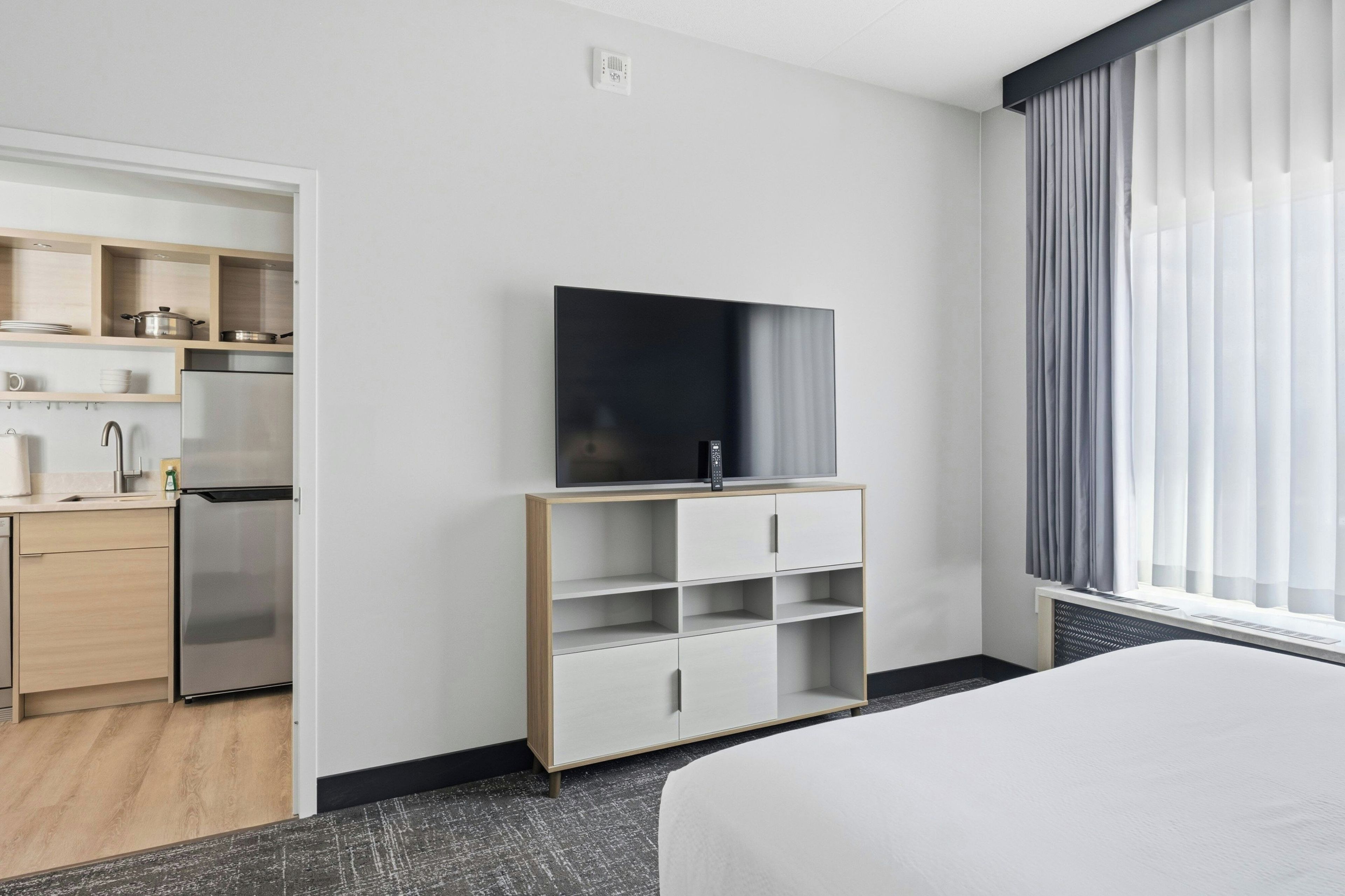 Een moderne hotelkamer met een bed, een televisie op een standaard, een kitchenette en een raam met gordijnen.