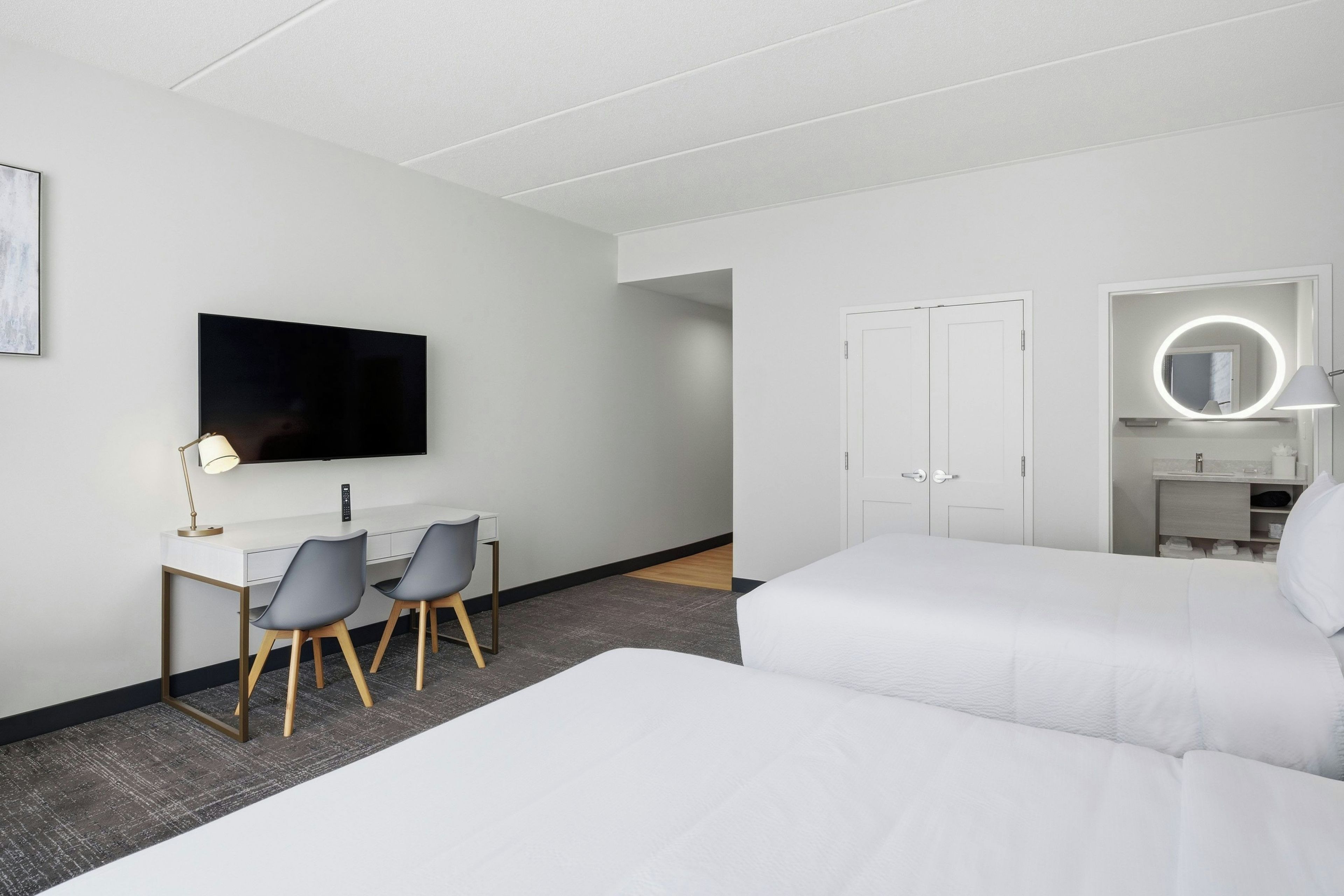 A hotel room with a bed, a desk with two chairs, a TV, a door, and a bathroom with a mirror.