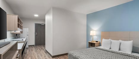 A hotel room with a bed, bedside table, lamp, and a kitchenette area.