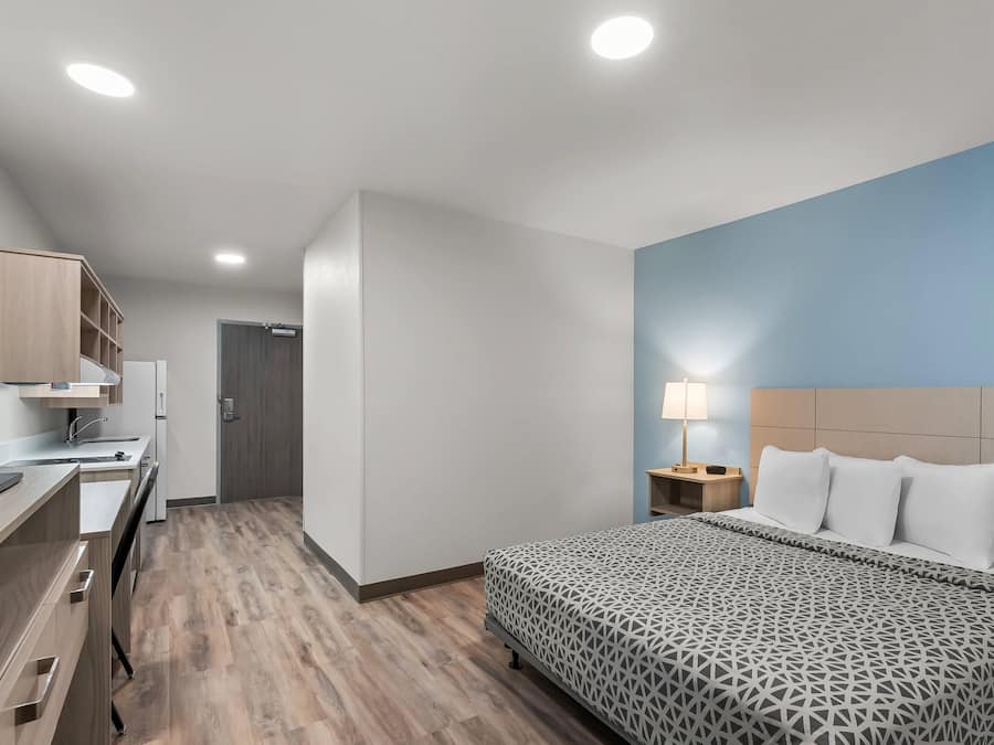 A hotel room with a bed, bedside table, lamp, and a kitchenette area.