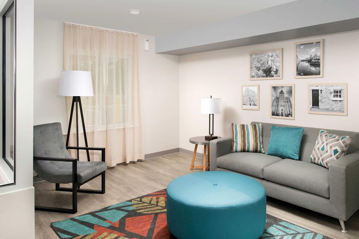 A modern living room with a grey sofa, a blue ottoman, a black chair, a floor lamp, and a patterned rug.