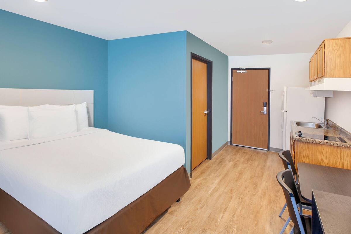 A hotel room with a bed, a desk, a chair, a kitchenette, and a door.