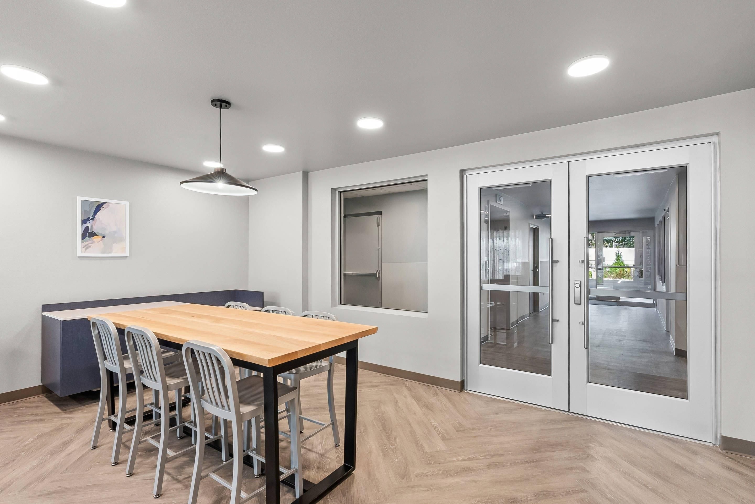 Un espace de salle à manger moderne avec une table en bois, des chaises en métal, des spots encastrés au plafond et une vue à travers une porte vitrée.