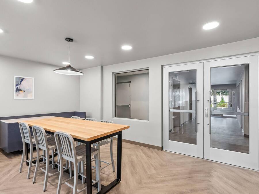 Un espace de salle à manger moderne avec une table en bois, des chaises en métal, des spots encastrés au plafond et une vue à travers une porte vitrée.