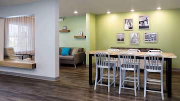 Un comedor moderno con una mesa de madera y sillas metálicas, un sofá con un cojín azul, y una ventana con cortinas de gasa.