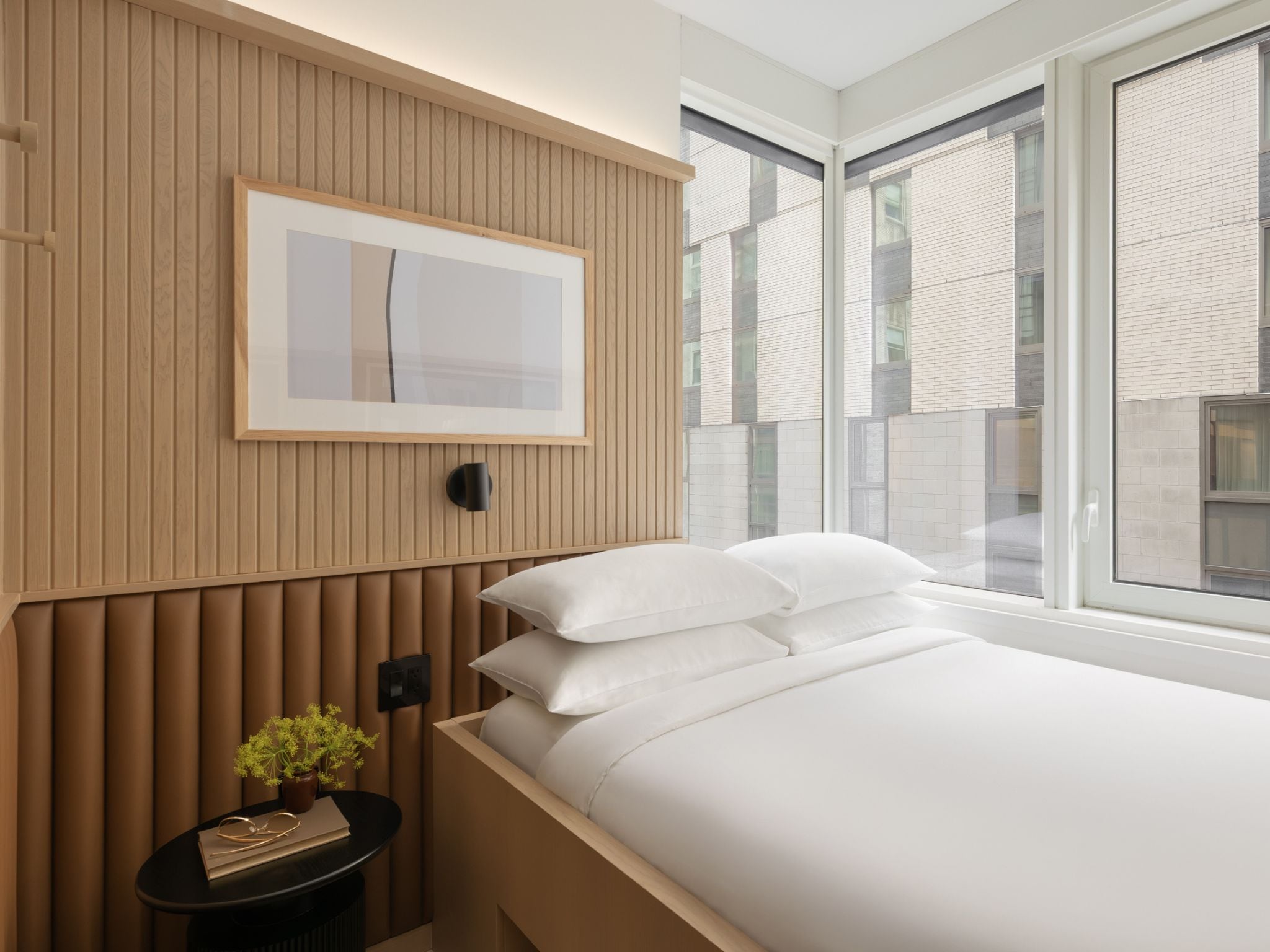 A modern bedroom with a bed, pillows, a bedside table with a plant, a framed picture on the wall, and large windows.