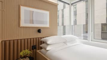 A modern bedroom with a bed, pillows, a bedside table with a plant, a framed picture on the wall, and large windows.