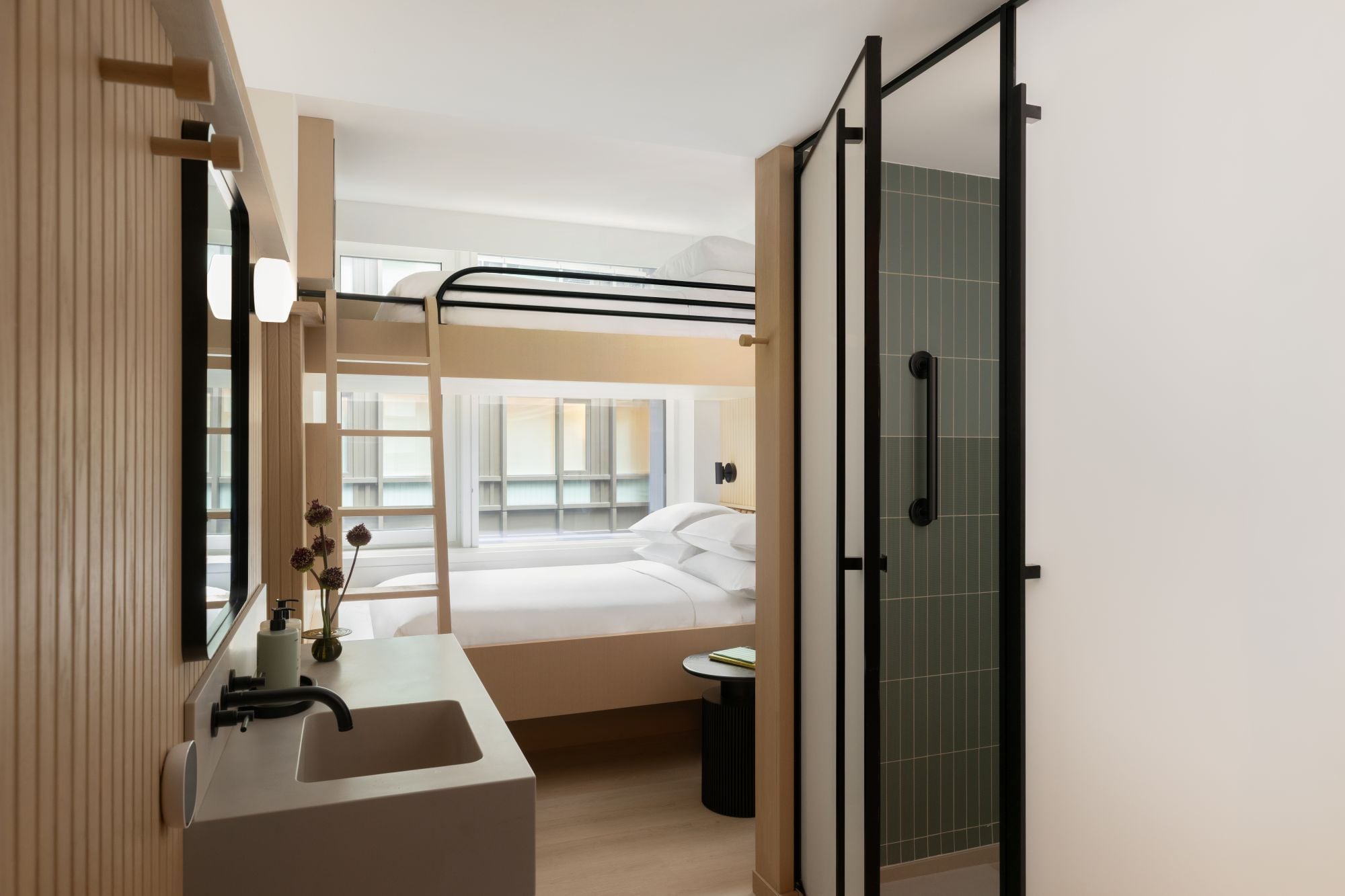 A modern hotel room with a bunk bed, a bathroom with a sink and mirror, and a glass shower enclosure.