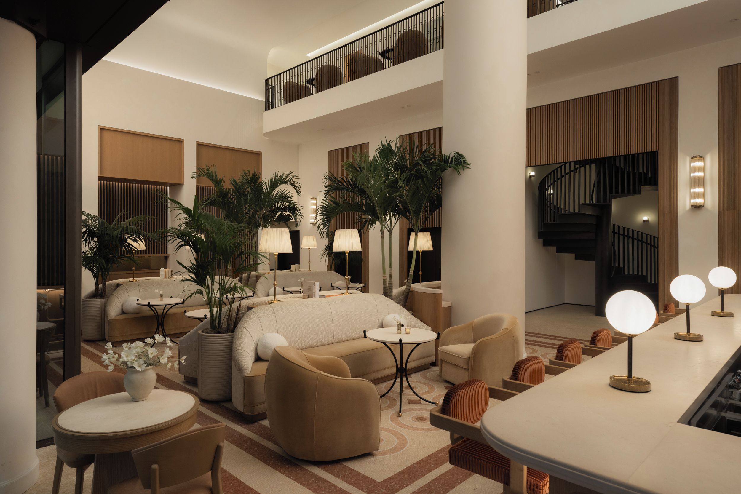 A spacious hotel lobby with a high ceiling, a central seating area with sofas and chairs, a staircase, and a reception desk with a bar.