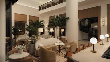 A spacious hotel lobby with a high ceiling, a central seating area with sofas and chairs, a staircase, and a reception desk with a bar.