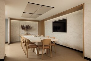 Un salón de conferencias con una mesa larga, sillas, un jarrón con plumas de pampa y un televisor en la pared.