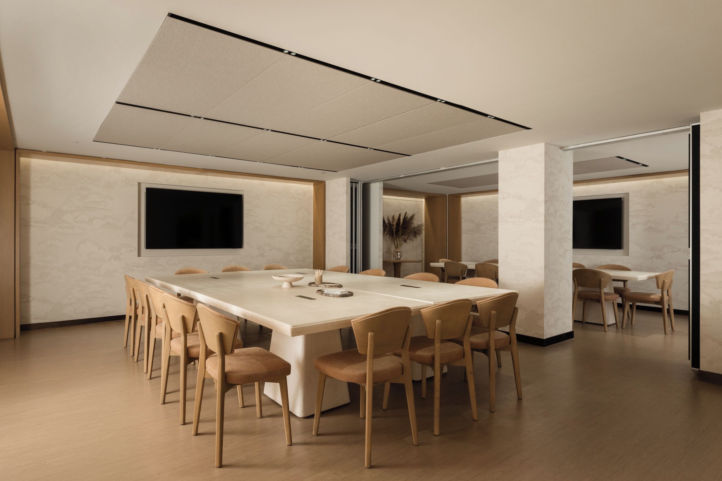 A conference room with a long table, multiple chairs, a vase with dried plants, and a large screen on the wall.